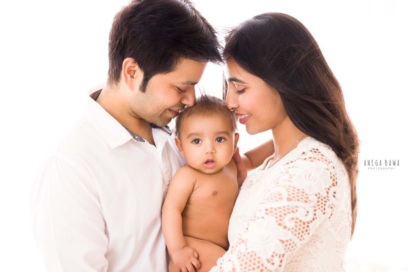 Captivating family photoshoot by Anega Bawa Photography, the esteemed photographer in Delhi and Gurgaon, capturing the tender moments of Mom, Dad, and their adorable 3, 4, 5, 6-month-old son in a kissing pose against a timeless white backdrop.