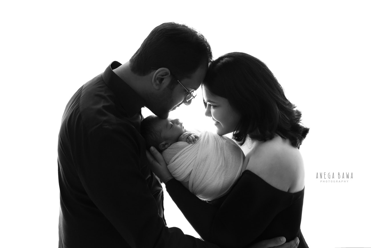 Captivating family photoshoot by Anega Bawa Photography, the esteemed photographer in Delhi and Gurgaon, portraying the endearing moments of Mom, Dad, and their beloved 3, 4, 5, 6-month-old son wrapped in white against a pristine white backdrop.