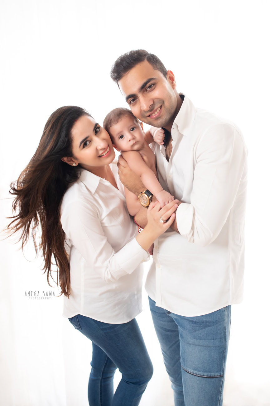 Captivating family photoshoot by Anega Bawa Photography, the esteemed photographer in Delhi and Gurgaon, featuring Mom and Dad dressed in white shirts and blue denim, along with their adorable 3, 4, 5, 6-month-old son against a clean white backdrop.
