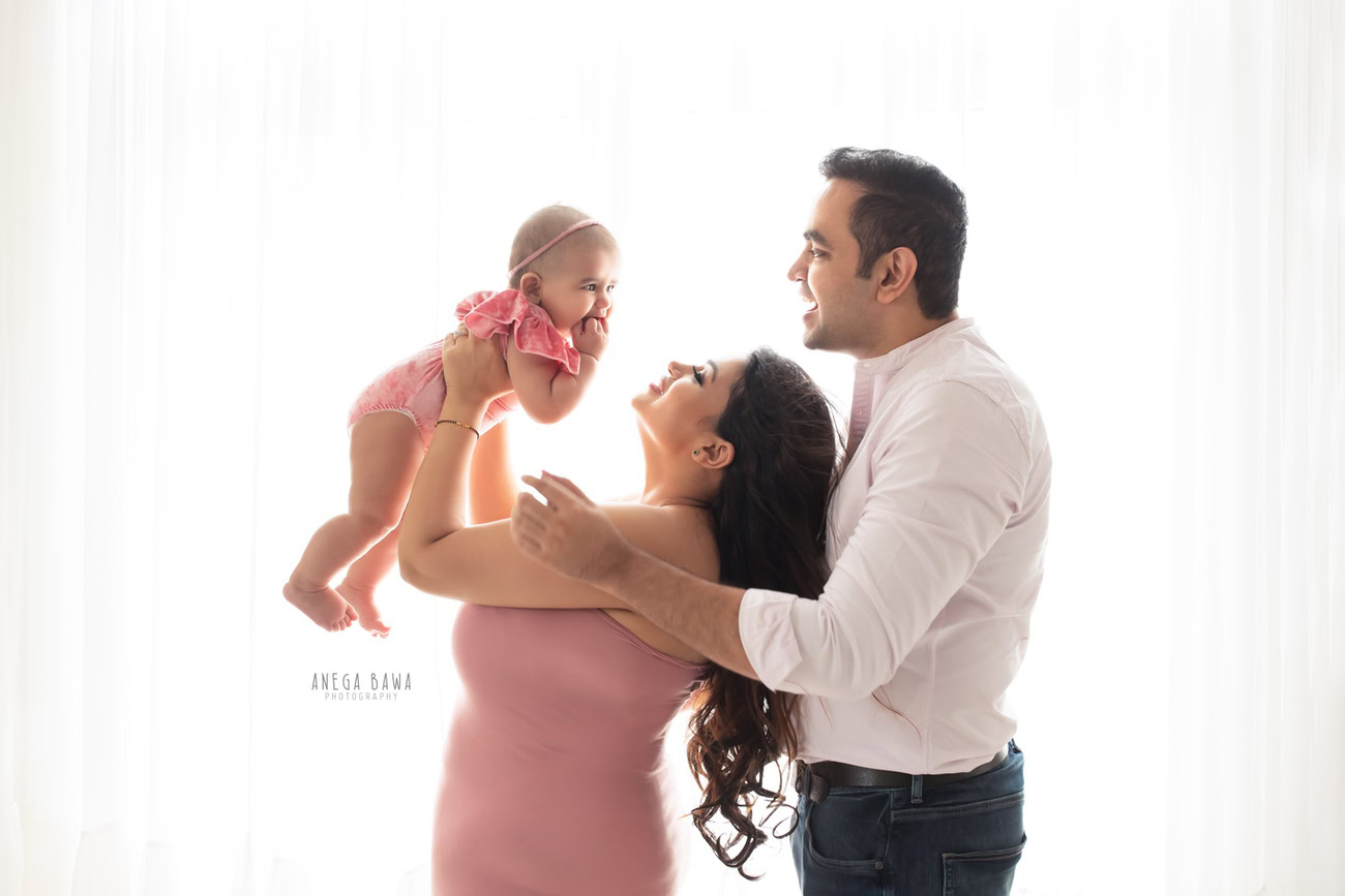 Heartwarming family photoshoot by Anega Bawa Photography, the renowned photographer in Delhi and Gurgaon, featuring a precious 3, 4, 5, 6-month-old baby girl with Mom and Dad against a pure white backdrop.