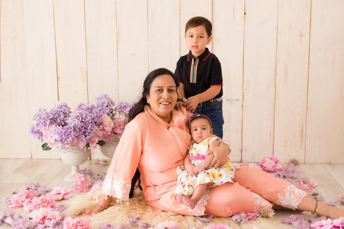 Captivating family photoshoot by Anega Bawa Photography, esteemed in Delhi and Gurgaon, featuring the heartwarming bond between Mom, daughter, and sibling, sitting on a beige rug with flowers scattered on the floor, against a harmonious beige backdrop.