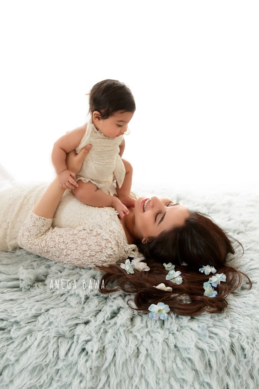 Captivating family photoshoot by Anega Bawa Photography, esteemed in Delhi and Gurgaon, capturing the precious moment of Mom and daughter sitting on Mom's tummy, adorned with a grey rug, against a serene white backdrop.
