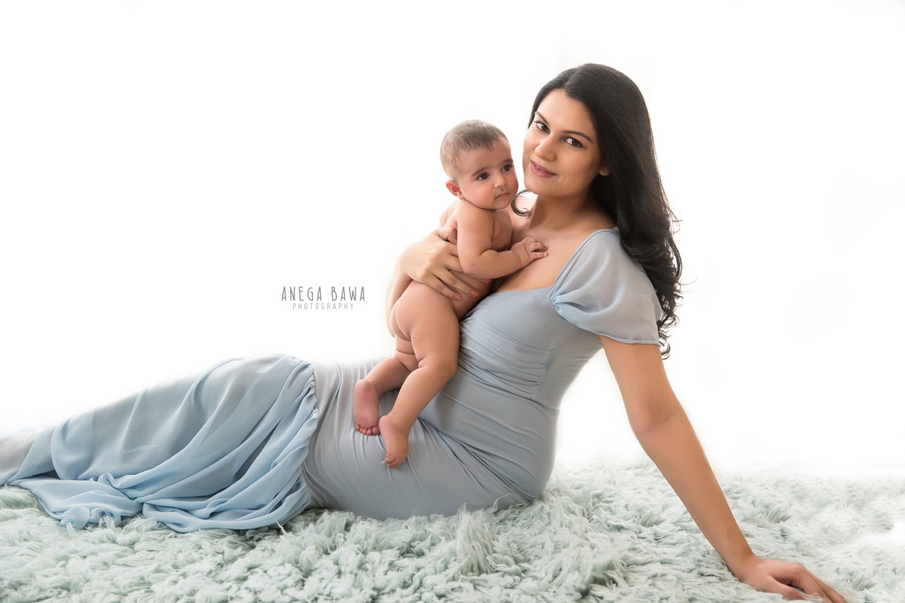 Captivating family photoshoot by Anega Bawa Photography, acclaimed in Delhi and Gurgaon, showcasing Mom in a sky-blue gown and her son sitting on a grey rug against a pristine white backdrop.