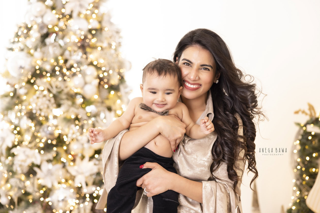 Captivating family photoshoot by Anega Bawa Photography, recognized in Delhi and Gurgaon, showcasing Mom and son with a cute bow, beside a festive Christmas tree, against a pristine white backdrop.