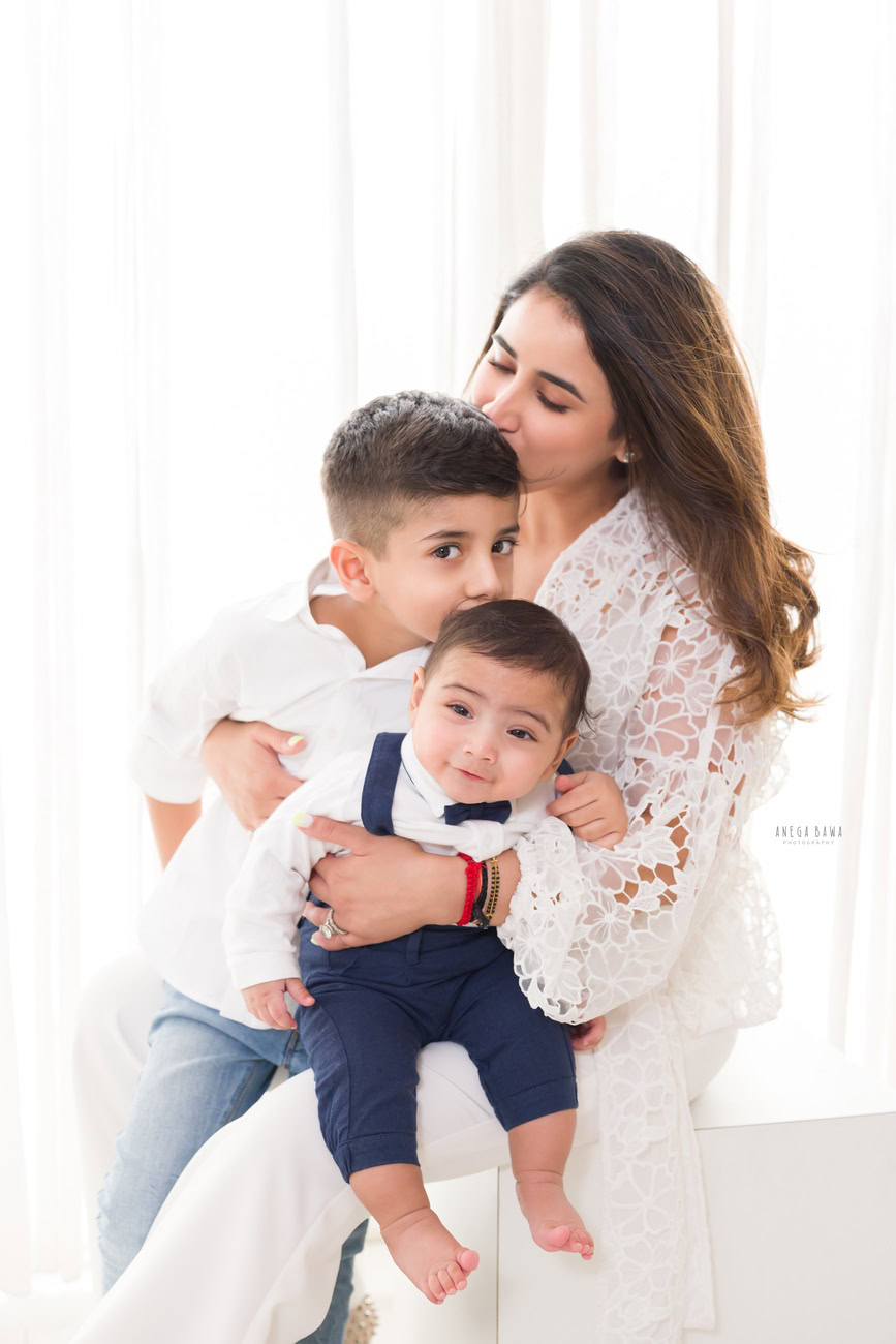 Capture the heartwarming bond between a mother, son, and sibling in a tender kissing pose against a pristine white backdrop. Book your session with Anega Bawa Photography in Delhi and Gurgaon for timeless family portraits.