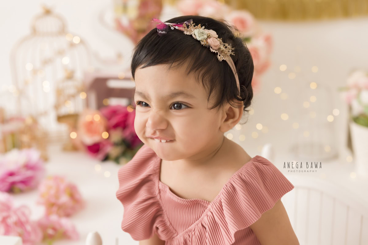 In this delightful toddler photoshoot captured by Anega Bawa in Delhi, Gurgaon, and Noida, a charming 12-month-old girl strikes a playful pose amidst a whimsical scene. Surrounded by enchanting castles, twinkling fairy lights, and delicate flowers, she exudes innocence and joy. Each image captures her mischievous spirit and the magic of childhood, creating timeless memories for her family to treasure.