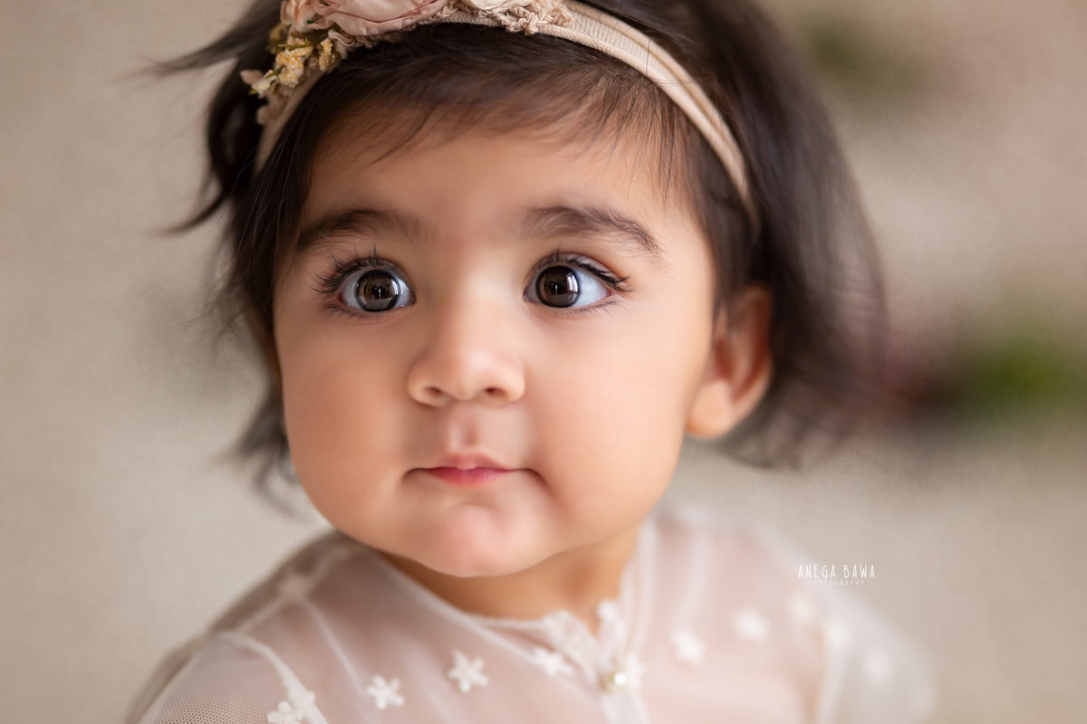 In this enchanting portrait by Anega Bawa, a 12-month-old girl captivates with her big twinkling eyes against a pristine white backdrop. With expert photography skills, Anega Bawa beautifully captures the innocence and wonder of this young child, creating a timeless image that celebrates the joy of childhood. This captivating photograph is sure to be cherished by the family for years to come.
