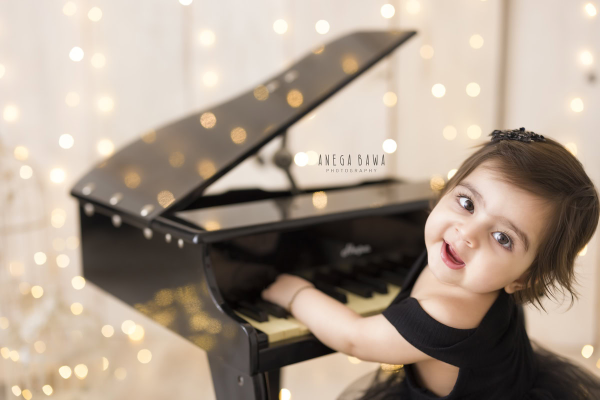 Girl in a charming black dress playing the piano amidst twinkling fairy lights against a serene beige backdrop, marking her journey from 12 months to 1 year old during her first birthday photoshoot in Delhi by Anega Bawa, Gurgaon, Noida.