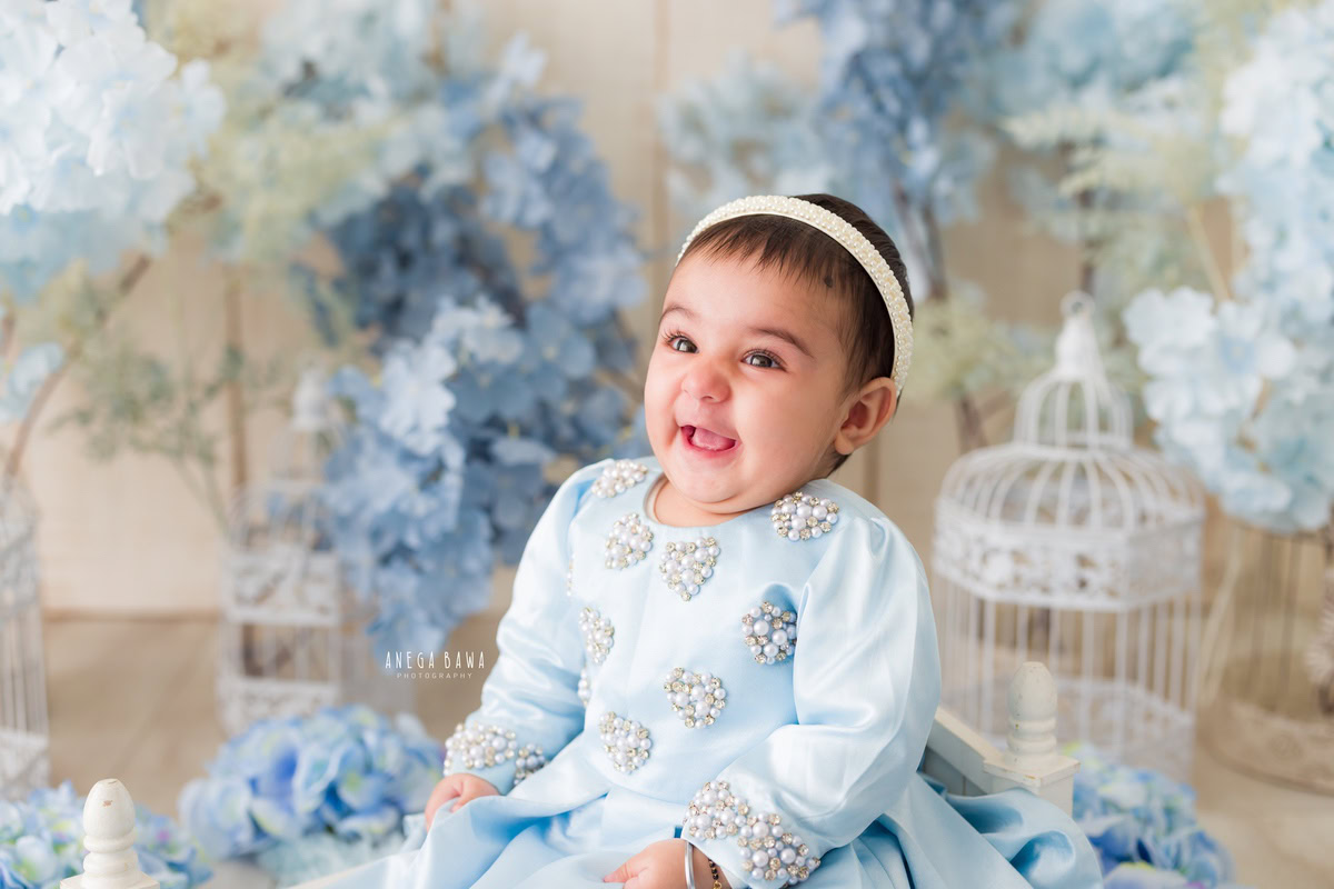 In this delightful toddler photoshoot by Anega Bawa in Delhi, Gurgaon, and Noida, a charming 12-month-old girl captivates in a lovely blue dress. Surrounded by whimsical white castles and delicate blue flowers against a serene beige backdrop, she exudes an aura of innocence and wonder. Each photograph beautifully captures her playful spirit and curiosity at this tender age, creating cherished memories for her family to treasure for years to come.