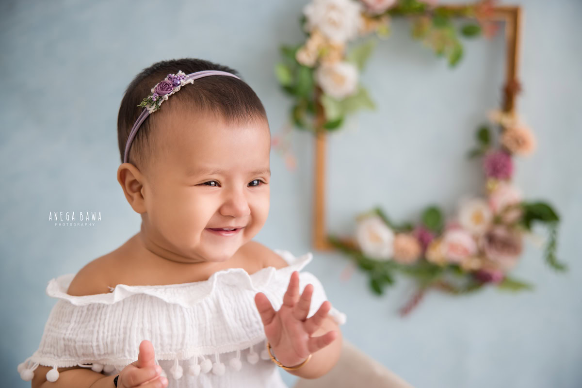 In this endearing sitter photoshoot by Anega Bawa in Delhi, Gurgaon, a delightful 12-month-old girl is captured in a heartwarming clapping pose. Against a serene blue backdrop, she exudes joy and innocence, her tiny hands clapping with enthusiasm. Framed by a wooden floral frame on the wall, this moment is a precious celebration of childhood captured for eternity.