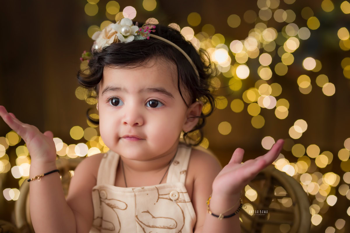 Celebrate the pure joy of your little princess with Anega Bawa's sitter photography in Delhi & Gurgaon. In this enchanting session, your 12-month to 1-year-old girl strikes a pose, clapping gleefully amidst twinkling fairy lights against a warm brown backdrop. Each captured moment is a testament to her innocence and happiness, creating cherished memories for years to come.