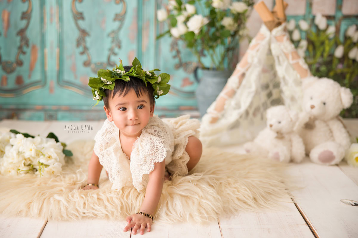 In this charming sitter photoshoot by Anega Bawa in Delhi, Gurgaon, a delightful 12-month-old girl is captured in a playful moment as she crawls on a soft beige rug. Against a backdrop of lush green, she explores her surroundings with curiosity and wonder. Adorned with a tiara band, she looks every bit the princess of her own little kingdom.
