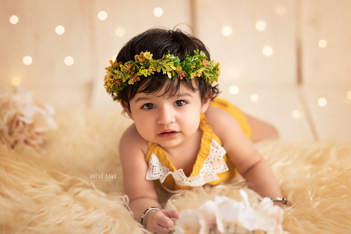 Embark on a magical journey with your little princess through a captivating photoshoot by Anega Bawa Photography in Delhi and Gurgaon. As she crawls gracefully on a plush beige rug, adorned with a delicate tiara band, surrounded by twinkling fairy lights against a backdrop of blooming florals, her innocence and beauty shine through at 12 months to 1 year old, creating enchanting memories to cherish forever.
