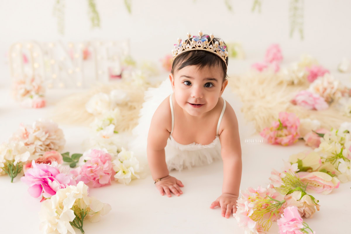 Witness the adorable milestones of your little one with Anega Bawa's sitter photography in Delhi & Gurgaon. In this captivating session, your 12-month to 1-year-old girl explores the world around her, crawling gracefully with a cute headband amidst scattered flowers, all against a pristine white backdrop. Every moment captured is a testament to her blossoming curiosity and charm, creating timeless memories to cherish forever.