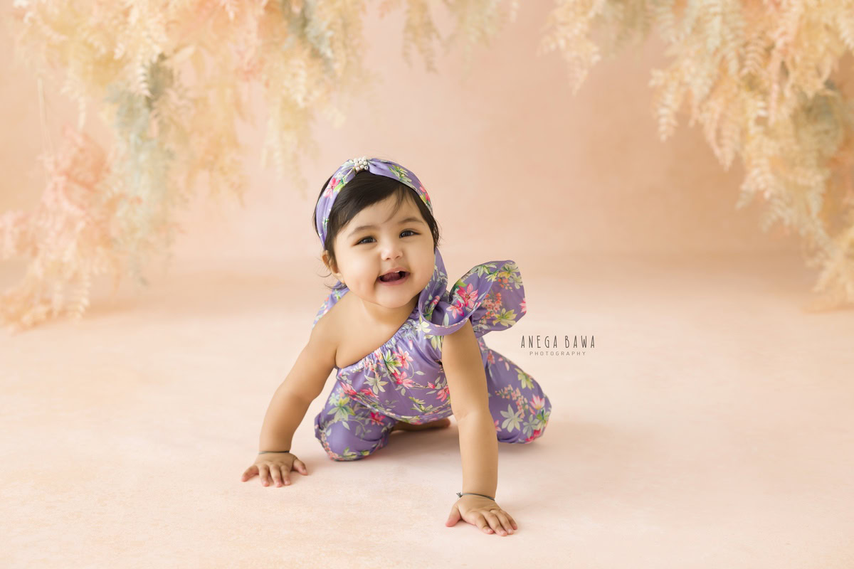 Captured during a baby pre-birthday photoshoot in Delhi, Anega Bawa, Gurgaon, and Noida, this 12-month-old girl crawls gracefully in a floral jumpsuit against a charming leafy peach backdrop.