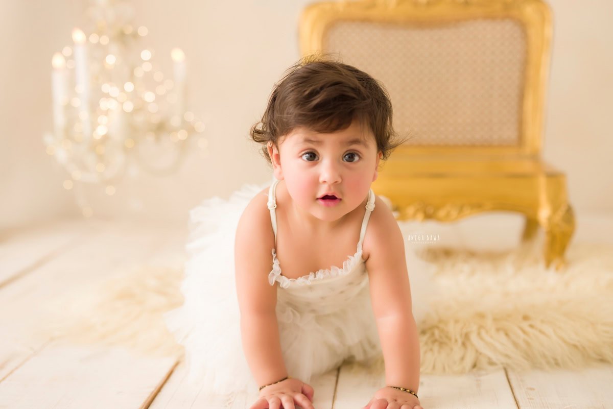 In this delightful capture from a baby pre-birthday photoshoot in Delhi, Anega Bawa, Gurgaon, and Noida, a 12-month-old girl crawls on a beige rug, adorned with golden lights and surrounded by the glow of candlelight. The scene is set against a serene beige backdrop, complementing the golden chair and candle frame beautifully.
