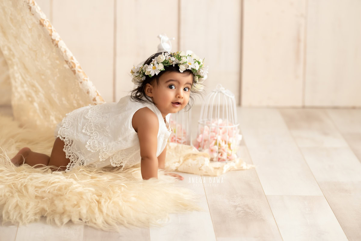 Witness your little princess's delightful exploration with Anega Bawa Photography in Delhi and Gurgaon. Adorned with a charming tiara band, she crawls gracefully on a beige rug, surrounded by white castles against a serene beige backdrop. Capture these endearing moments of your 12-month to 1-year-old and treasure them forever.