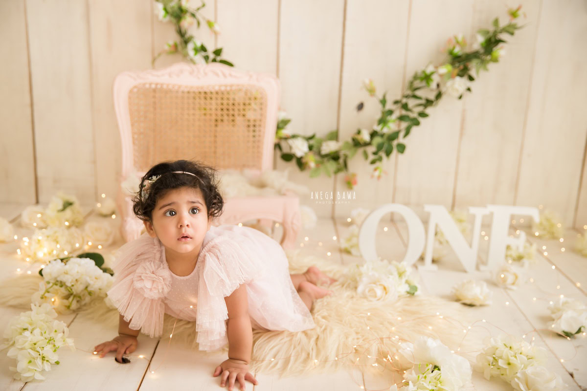 In this enchanting sitter photoshoot by Anega Bawa in Delhi, Gurgaon, a sweet 12-month-old girl is captured in a moment of exploration. Crawling on a soft beige rug adorned with delicate white flowers, she exudes innocence and curiosity. Against a backdrop of beige, a floral fringe on the wall adds a touch of whimsy to the scene, enhancing the overall charm of the photo.