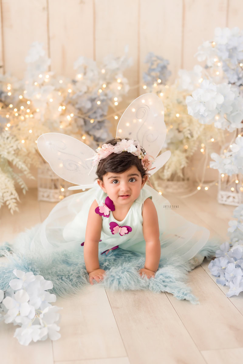 In this sitter birthday photoshoot by Anega Bawa in Delhi, Gurgaon, or Noida, a 12-month-old girl adds charm to the scene as she crawls on a blue rug. Adorned with butterfly wings, she brings a playful and whimsical touch to the setup. The beige backdrop provides a neutral background, allowing the focus to remain on the adorable little crawler.