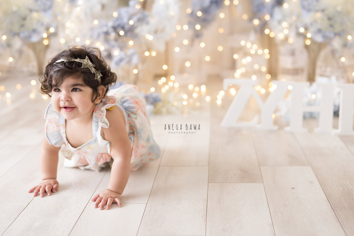 In this enchanting toddler photoshoot by Anega Bawa in Delhi, Gurgaon, and Noida, a sweet 12-month-old girl explores her surroundings with curiosity and joy. Crawling on the floor adorned with delicate blue and white flowers, she is surrounded by twinkling fairy lights that add a magical touch to the scene. Each moment captured in this photoshoot reflects the innocence and wonder of childhood, creating cherished memories for her family to treasure for years to come.