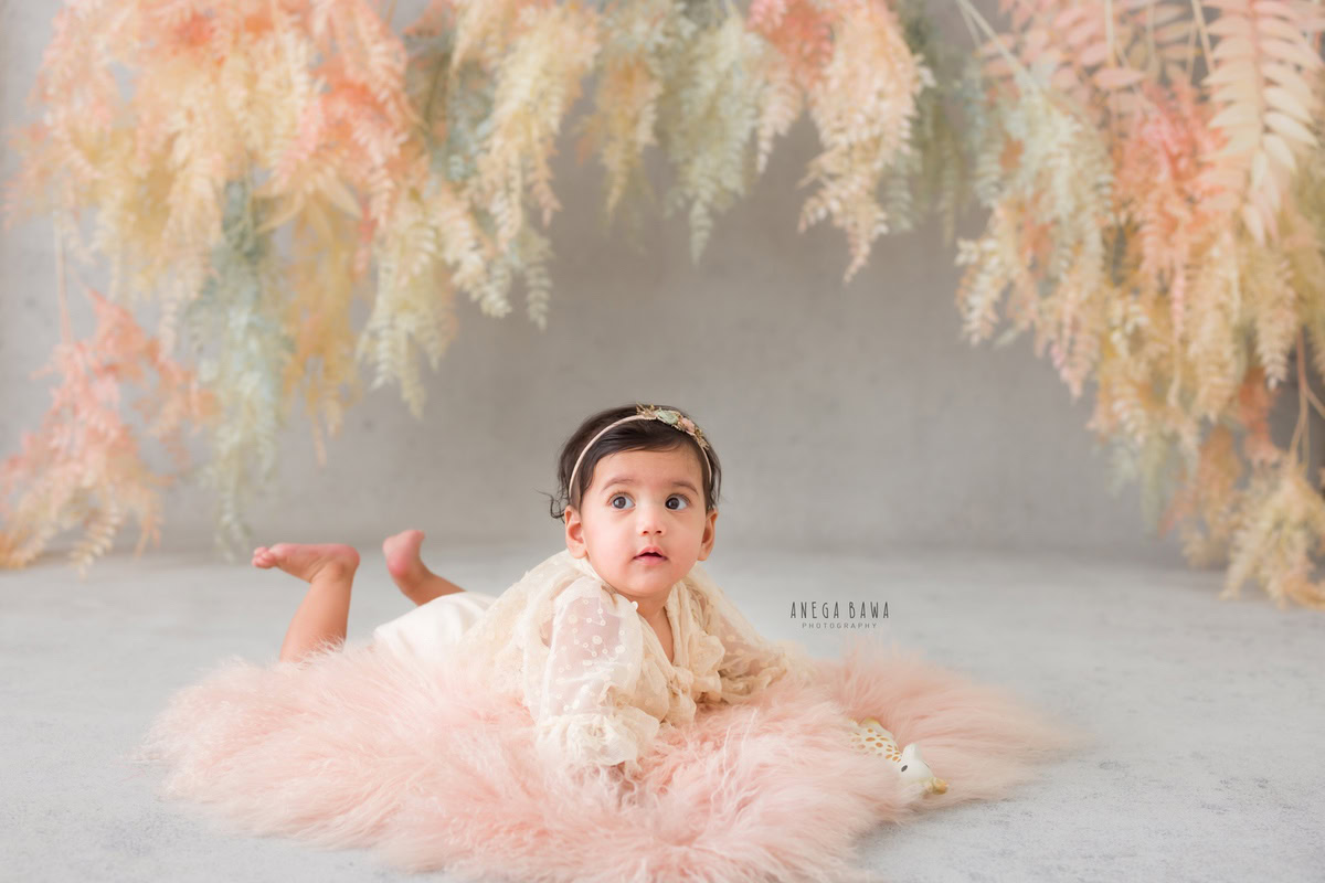 In this delightful toddler photoshoot captured by Anega Bawa in Delhi, Gurgaon, and Noida, a charming 12-month-old girl explores her world with curiosity and joy. Crawling on a soft pink rug against a backdrop of lush green leaves, she exudes innocence and wonder. Each moment captured in this photoshoot is a testament to the beauty and magic of childhood, creating cherished memories for her family to treasure forever.