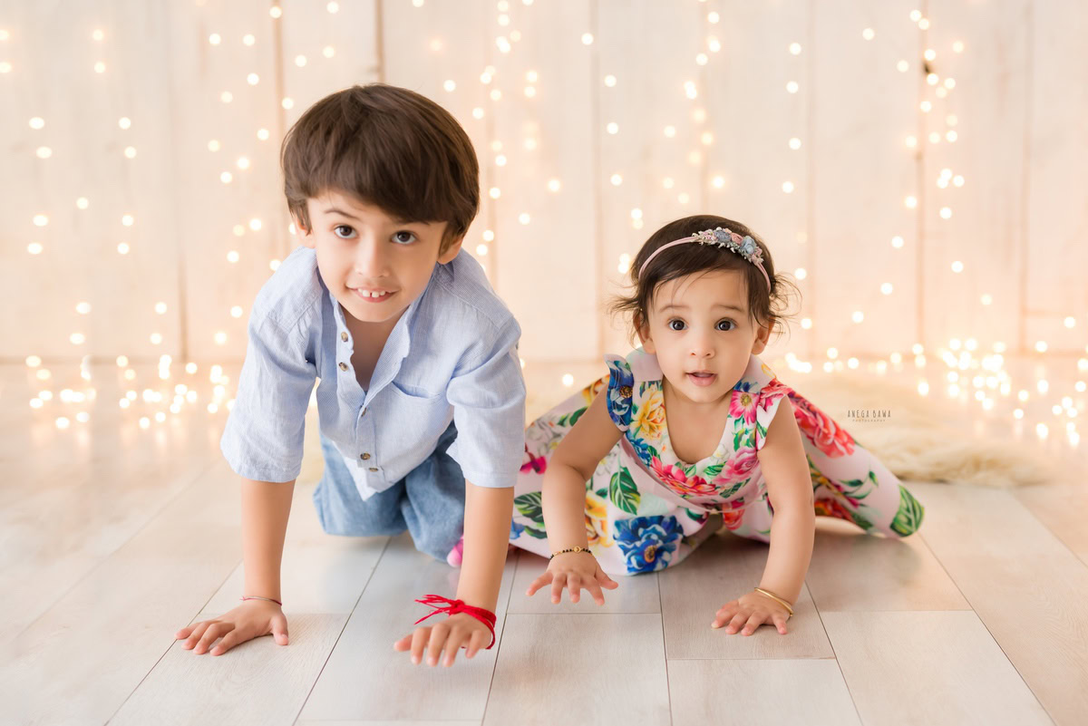 In this enchanting moment captured during a baby pre-birthday photoshoot in Delhi, Anega Bawa, Gurgaon, and Noida, a 12-month-old girl crawls alongside her sibling on a beige backdrop illuminated by twinkling fairy lights.