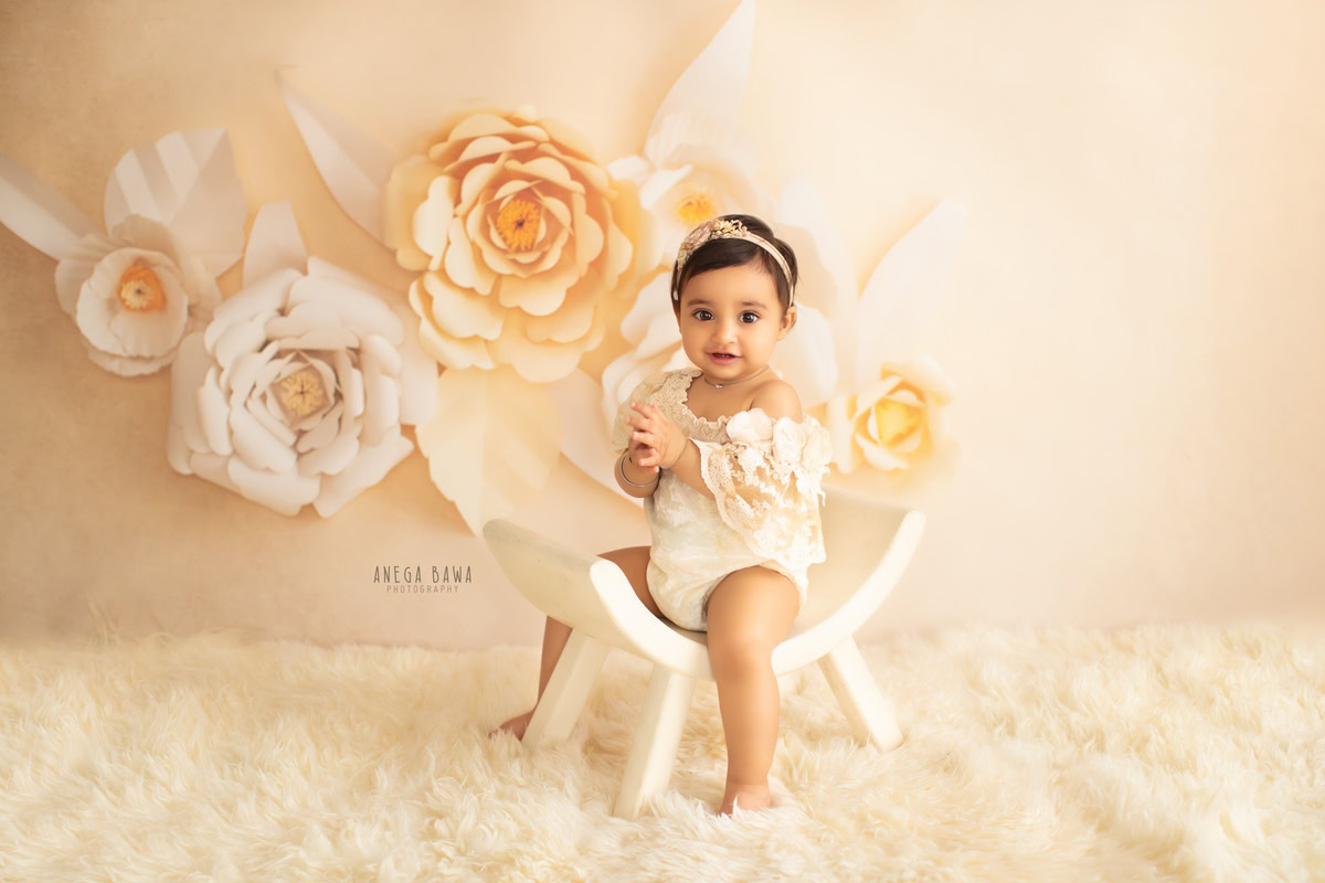 In this charming portrait by Anega Bawa, a 12-month-old girl sits gracefully on a curvy chair, adorned with a delicate beige rug, against a backdrop of vibrant floral patterns. With expert photography skills, Anega Bawa captures the innocence and beauty of childhood in this delightful image. This enchanting photograph is a testament to the skill and artistry of the photographer, creating a timeless keepsake for the family to treasure for years to come.