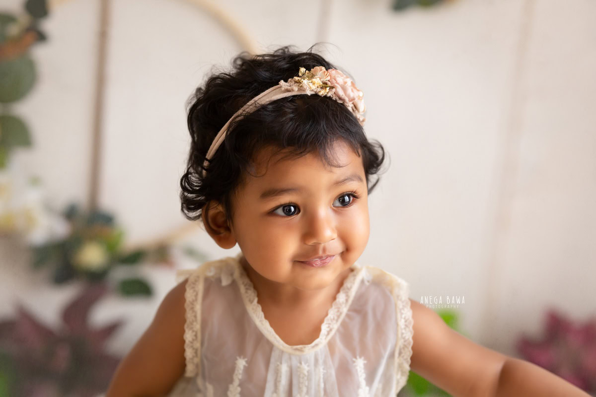 At 11 months, this adorable girl steals the show with her cute headband against a white backdrop. The leafy fringe on the wall adds a touch of natural charm to the scene.