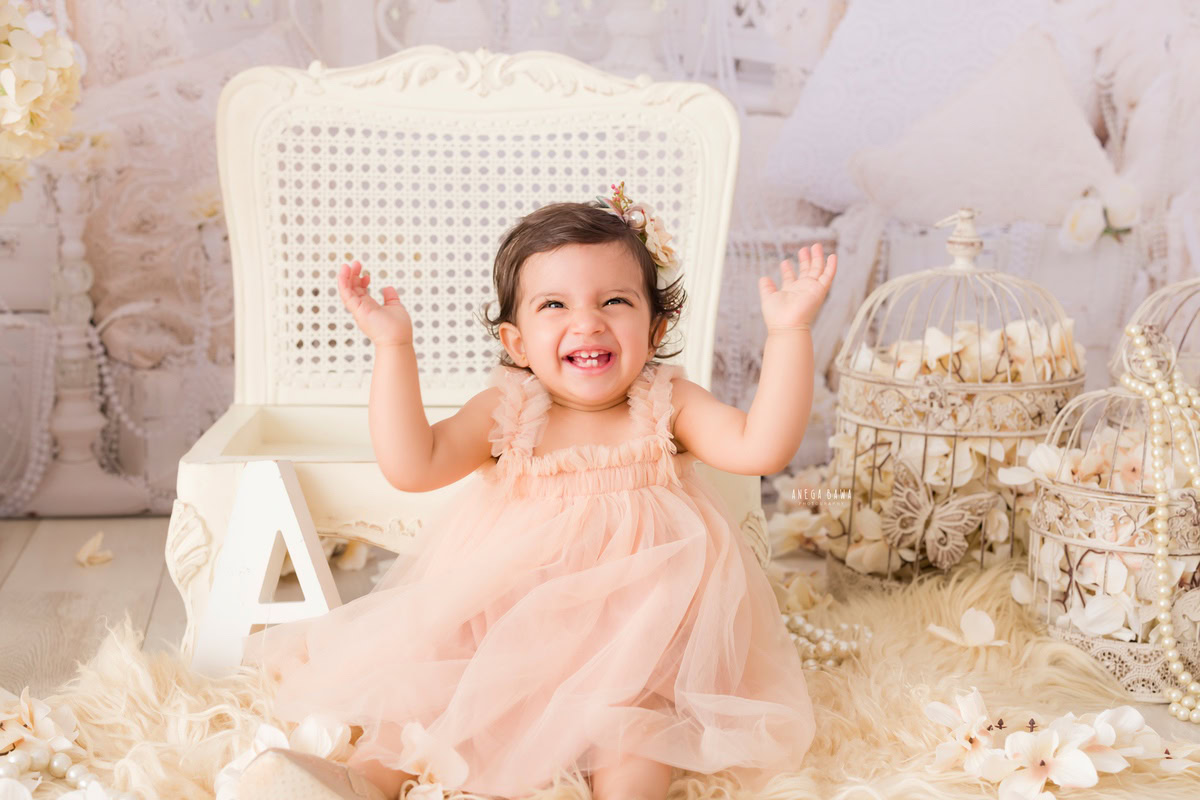 Celebrate the joyous laughter of your little girl with Anega Bawa's sitter photography in Delhi & Gurgaon. In this delightful session, your 11-month to 1-year-old princess exudes pure happiness as she poses on a white wooden chair, surrounded by a cozy beige rug and whimsical white castles against a serene white backdrop. Each click captures her infectious laughter, creating timeless memories of her precious moments.