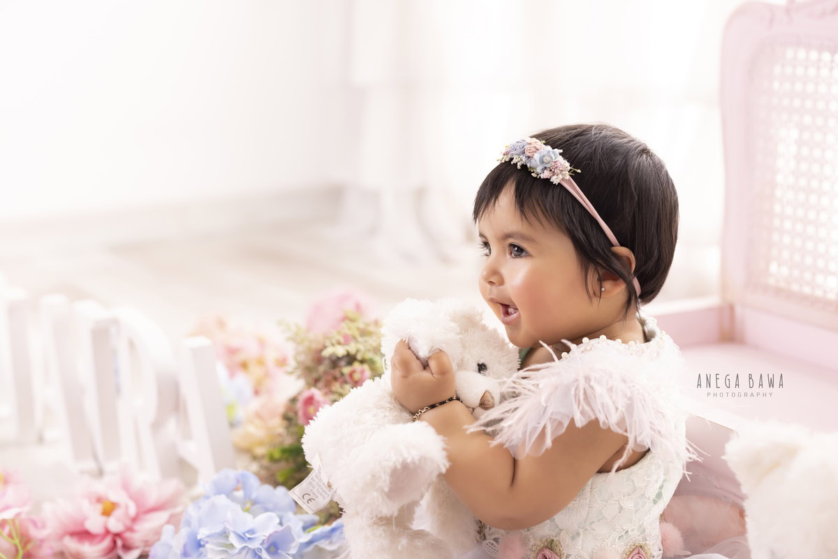 Girl adorned in a delicate feather dress, holding a teddy bear amidst pink and blue flowers against a pristine white backdrop, commemorating her journey from 11 months to 1 year old during her first birthday photoshoot in Delhi by Anega Bawa, Gurgaon, Noida.