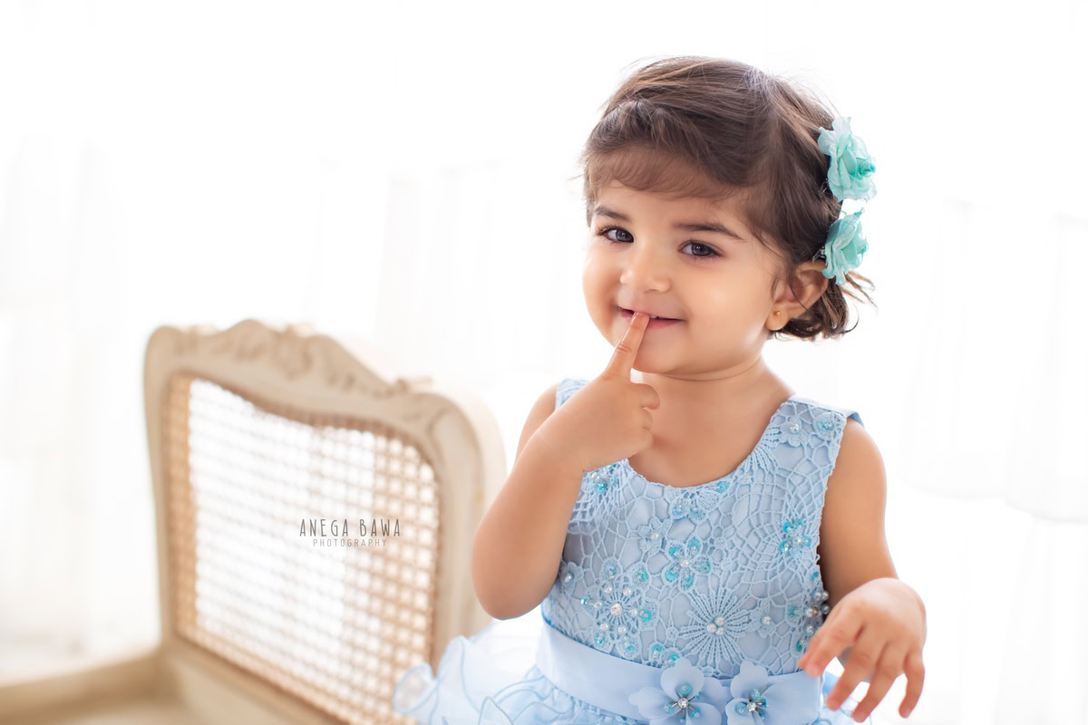 Celebrate your little one's blossoming personality with Anega Bawa Photography in Delhi and Gurgaon. Adorned with a delicate floral accessory in her hair, she sits gracefully on a white wooden chair against a pristine white backdrop. Capture the charm of your 11-month to 1-year-old in this enchanting photoshoot, creating timeless memories to cherish.