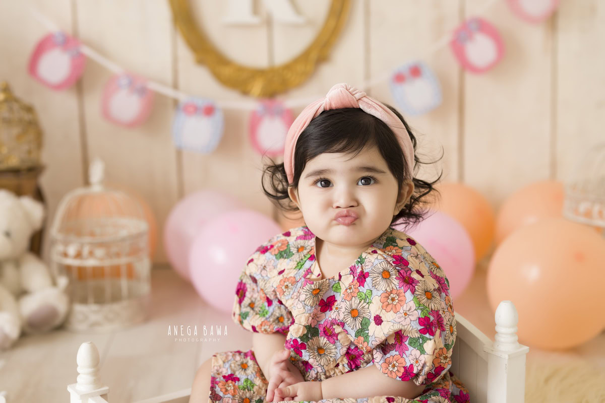 In this delightful toddler photoshoot by Anega Bawa in Delhi, Gurgaon, and Noida, a charming 11-month-old girl shines in a floral dress paired with a cute headband. Surrounded by balloons and teddy bears against a beige backdrop adorned with ivory castles, she exudes innocence and joy. Each frame captures her playful spirit and enchanting smile, creating precious memories for her family to cherish forever.