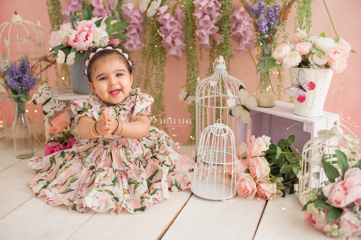 Captured during a sitter photo shoot in Delhi, Gurgaon by Anega Bawa, this adorable 11-month-old girl is dressed in a charming floral dress and headband. The scene unfolds against a pink backdrop, with fairy lights and white castles adding to the enchanting atmosphere.