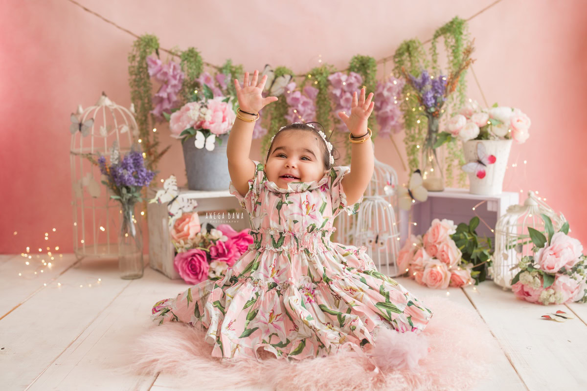 In this delightful image captured during a sitter photo shoot in Delhi, Gurgaon by Anega Bawa, a 11-month-old girl shines in a floral dress against a pink backdrop. The scene is further adorned with a floral fringe on the wall and a pink rug, adding to the charming ambiance.