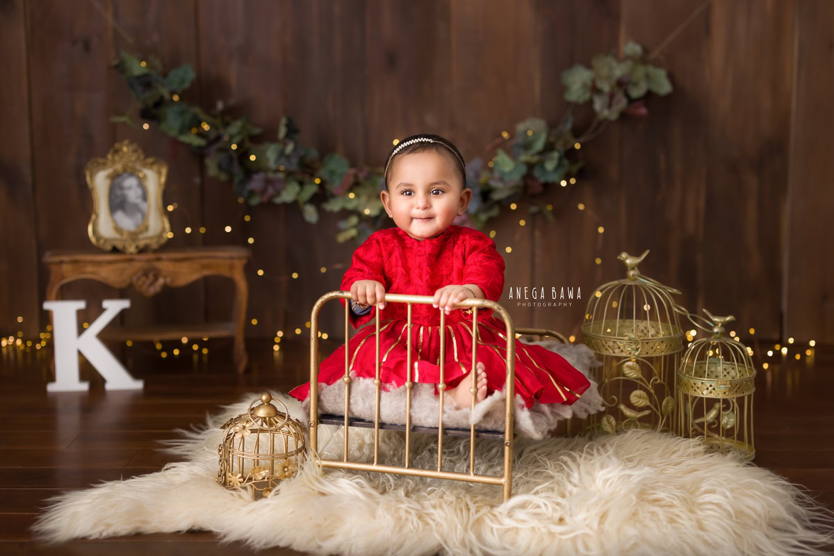 In this enchanting moment from a sitter photo shoot in Delhi, Gurgaon by Anega Bawa, a 11-month-old girl sits gracefully in a golden cot, accentuated by a beige rug. Against a brown backdrop, the scene is embellished with a fringe of leaves on the wall, illuminated by golden lights. Golden castles and an alphabet adorn the wall, adding a whimsical touch to the setting.