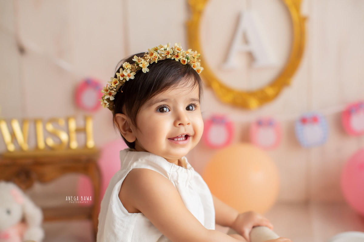 Let Anega Bawa Photography in Delhi and Gurgaon immortalize the innocence of your little princess. Adorned with a tiara band, she holds onto a chair beside an alphabet frame, set against a serene beige backdrop, creating timeless memories at 11 months to 1 year old.