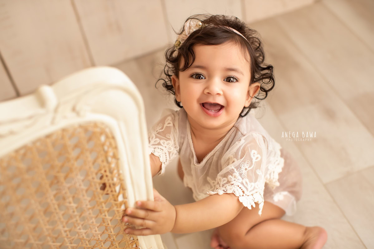 In this heartwarming capture by Anega Bawa, a radiant 11-month-old girl exudes joy as she holds onto a chair, showcasing a charming smile and adorned with a delightful headband. The skilled photographer perfectly encapsulates the innocence and happiness of childhood, creating a cherished memory for the family. This enchanting photograph is a testament to the photographer's talent and expertise, offering a timeless keepsake for the family to treasure forever.