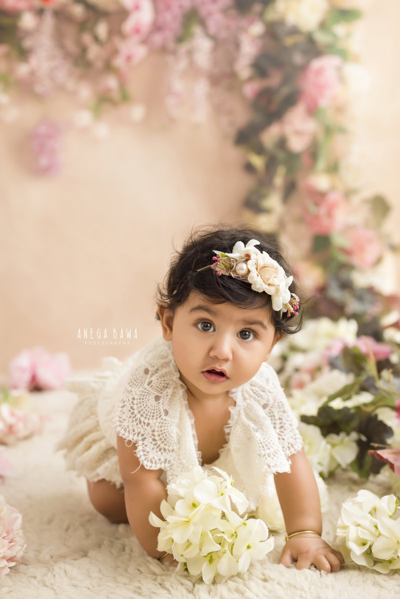 In this heartwarming toddler photoshoot captured by Anega Bawa in Delhi, Gurgaon, and Noida, a precious 11-month-old girl holds a delicate flower against a backdrop of soft beige. The floral fringe adds a touch of whimsy to the scene, perfectly complementing the girl's innocent charm. Each frame captures her beauty and innocence, creating timeless memories for her family to cherish for years to come.