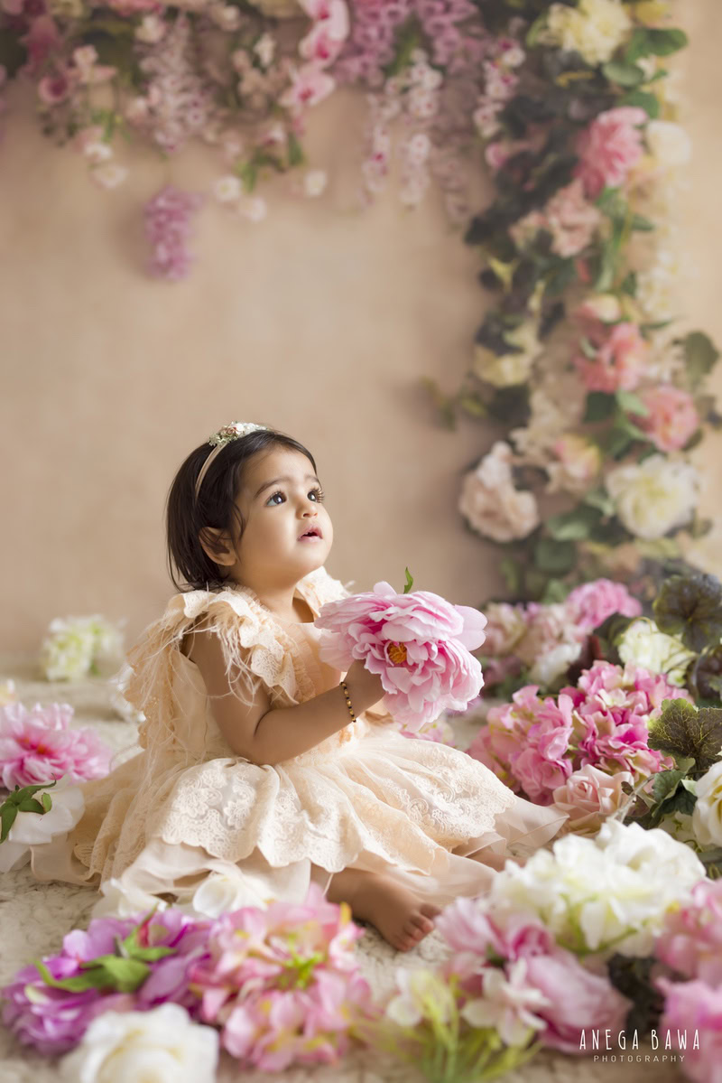 In this delightful toddler photoshoot by Anega Bawa in Delhi, Gurgaon, and Noida, a charming 11-month-old girl holds a lovely flower against a backdrop of soft peach. The floral fringes add a whimsical touch to the scene, enhancing the girl's natural beauty and innocence. Each photograph captures her adorable personality, creating cherished memories for her family.