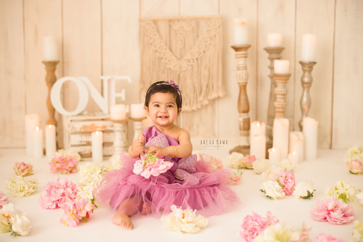 In this captivating frame captured during a sitter photo shoot in Delhi, Gurgaon by Anega Bawa, an 11-month-old girl exudes charm as she delicately holds flowers against a serene beige backdrop. The scene is adorned with flowers scattered on the floor, adding a touch of natural beauty to the setting.
