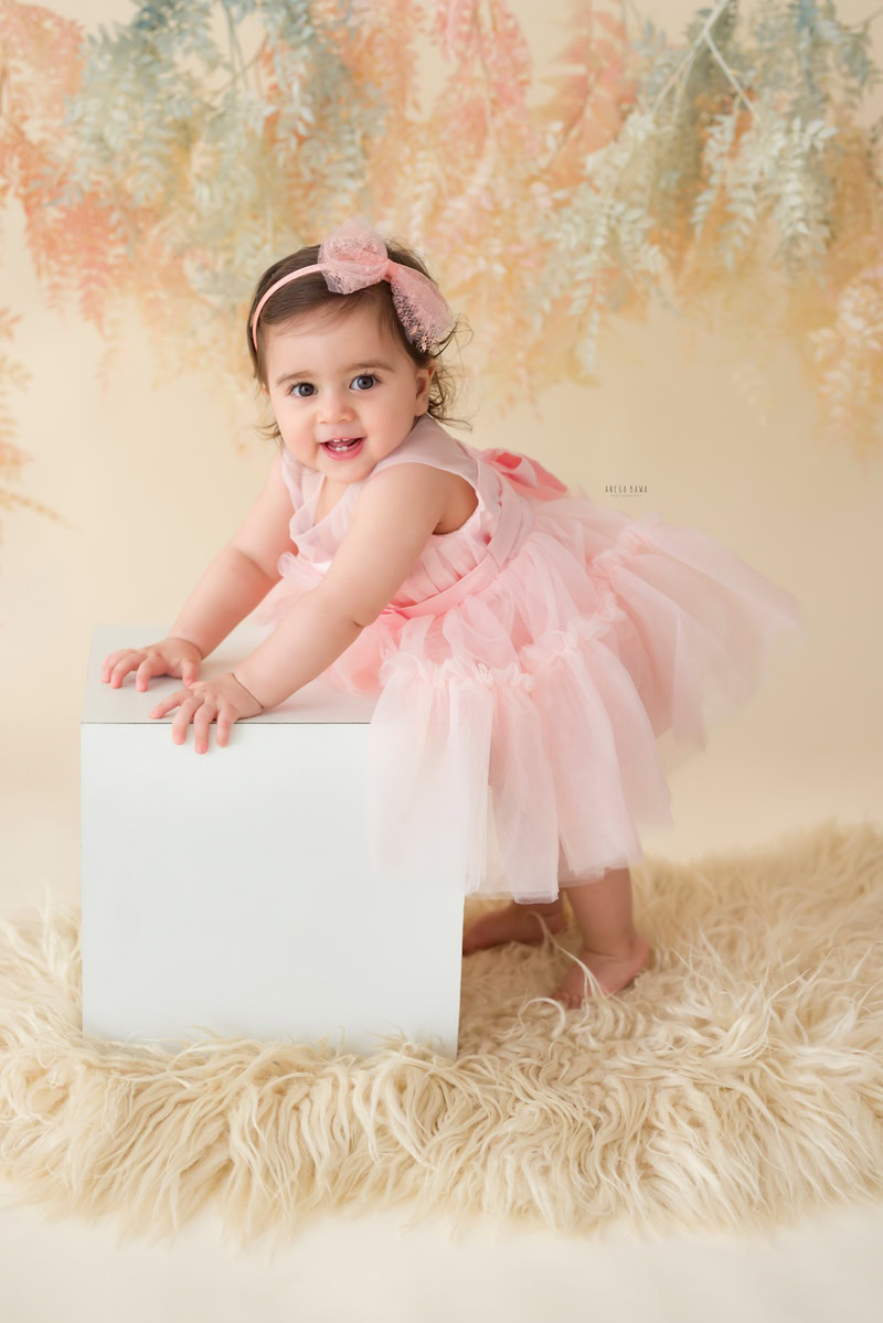 In this captivating toddler photoshoot by Anega Bawa in Delhi, Gurgaon, and Noida, an adorable 11-month-old girl steals the spotlight as she holds a stool with a pink headband adorning her hair. The beige rug provides a soft contrast against the lush leafy peach backdrop, creating a picturesque setting for capturing her precious moments. Each photograph reflects her innocent charm and playful spirit, making it a cherished memory for her family to treasure forever.