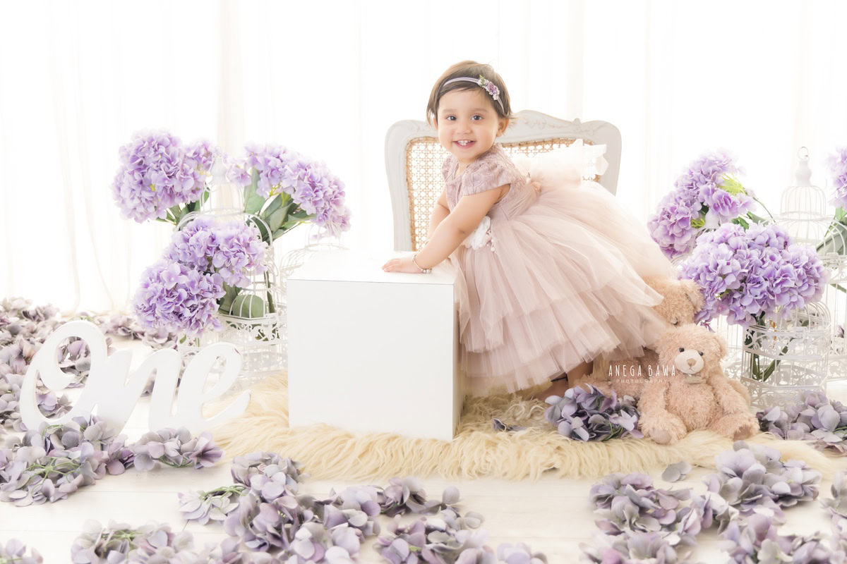 In this enchanting toddler photoshoot captured by Anega Bawa in Delhi, Gurgaon, and Noida, a delightful 11-month-old girl is portrayed holding a white stool, surrounded by a lavender bunch of flowers against a serene white backdrop. This charming scene beautifully encapsulates the innocence and grace of childhood, creating timeless memories for her family to cherish for years to come.