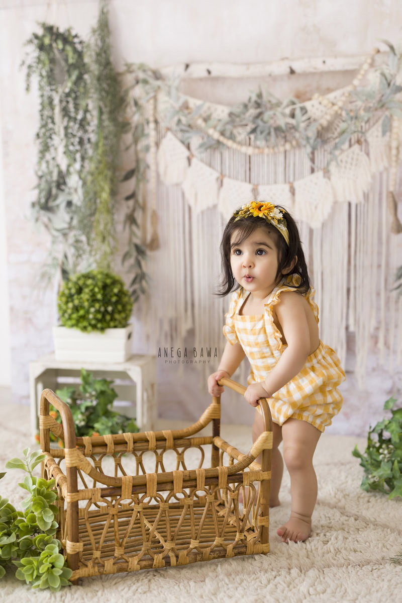 Girl holding a wooden cot adorned with a sunflower headband against a pristine white backdrop, with leafy bunches hanging on the wall, marking her journey from 11 months to 1 year old during her first birthday photoshoot in Delhi by Anega Bawa, Gurgaon, Noida.