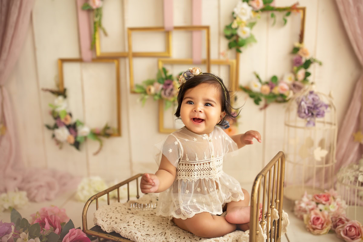 Step into a world of enchantment with Anega Bawa Photography in Delhi and Gurgaon. Your little princess rests in a golden cot surrounded by floral frames and castles amidst pink flowers, all against a captivating beige backdrop, capturing the magic of her 11 months to 1 year old journey.