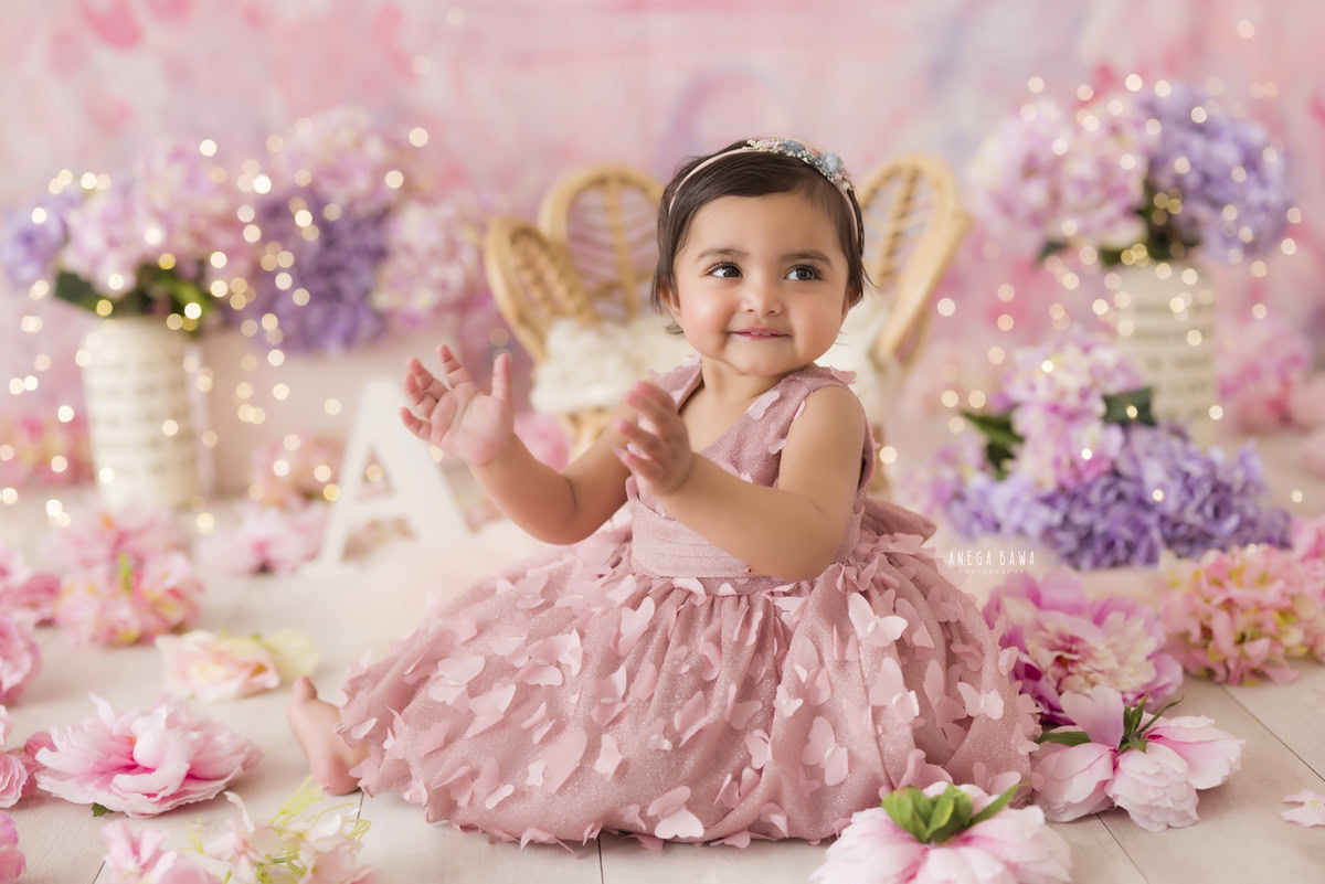 In this captivating toddler photoshoot by Anega Bawa in Delhi, Gurgaon, and Noida, a delightful 11-month-old girl is adorned in a pink dress, surrounded by a charming display of lavender and pink flowers illuminated by fairy lights against a soft pink backdrop. This enchanting scene captures the innocence and joy of childhood, creating treasured memories for her family to cherish for a lifetime.