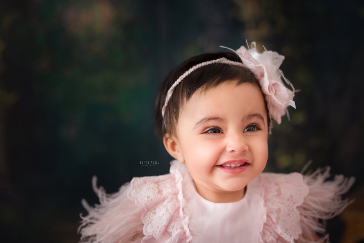 Indulge in the enchanting beauty of your little princess with Anega Bawa's sitter photography in Delhi & Gurgaon. Dressed in a charming pink feather dress and adorned with a matching headband, she radiates elegance against a warm brown backdrop. This delightful session captures the grace and innocence of your 11-month to 1-year-old daughter, preserving her precious moments in timeless photographs.