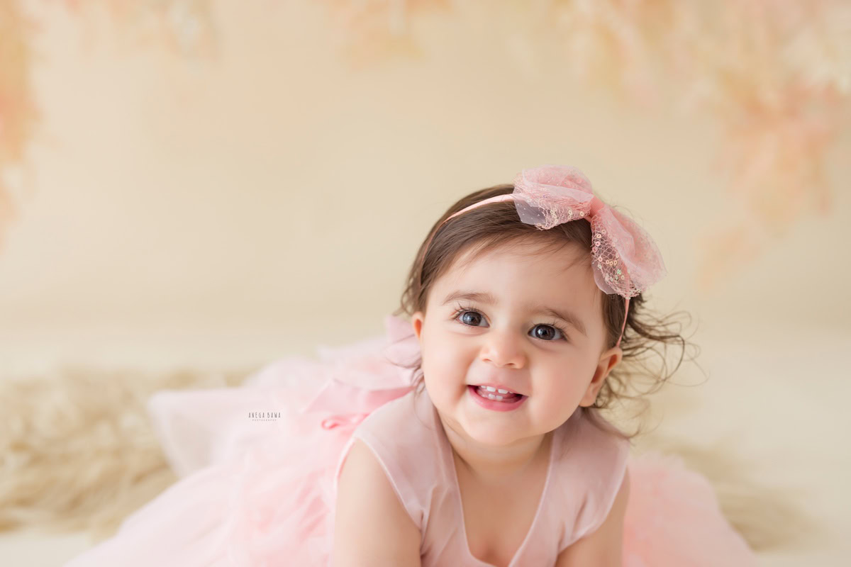 Capture the adorable charm of your little girl with Anega Bawa's sitter photography in Delhi & Gurgaon. Dressed in a sweet pink headband and seated on a cozy pink rug against a soft beige backdrop, she exudes innocence and warmth. This delightful session preserves the precious moments of your 11-month to 1-year-old daughter, creating timeless memories for you to cherish.