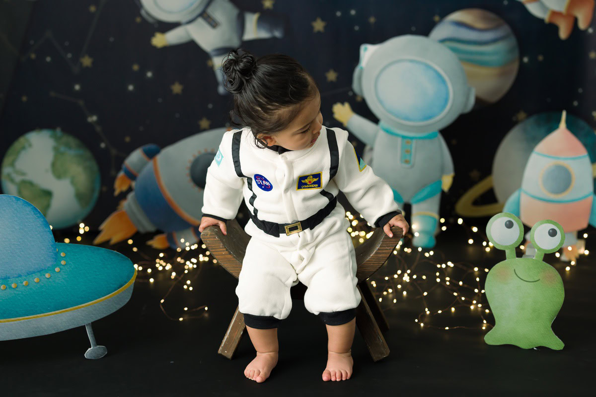 Delight in the cosmic charm of your little one with Anega Bawa's sitter photography in Delhi & Gurgaon. Adorned in a space-themed outfit, she sits gracefully on a curvy chair against a stellar backdrop, illuminated by twinkling fairy lights. This enchanting session captures the celestial spirit of your 11-month to 1-year-old daughter, creating timeless memories to treasure forever.