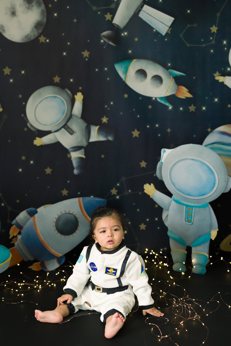 In this magical toddler photoshoot by Anega Bawa in Delhi, Gurgaon, and Noida, an adventurous 11-month-old girl is dressed in a charming space outfit, surrounded by a captivating space backdrop adorned with twinkling fairy lights. This whimsical scene captures the wonder and imagination of childhood, creating cherished memories for her family to treasure forever.