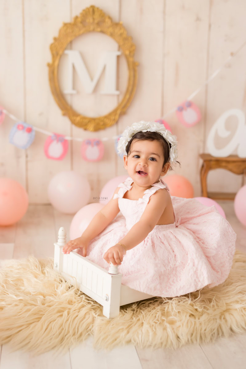In this delightful toddler photoshoot by Anega Bawa in Delhi, Gurgaon, and Noida, a precious 10-month-old girl is nestled in a cozy white cot, wearing an adorable headband. Behind her, a golden frame on the wall adds a touch of elegance to the scene, complemented by a soft beige rug and backdrop. This enchanting setup captures the innocence and beauty of infancy, creating timeless memories for her family to cherish.